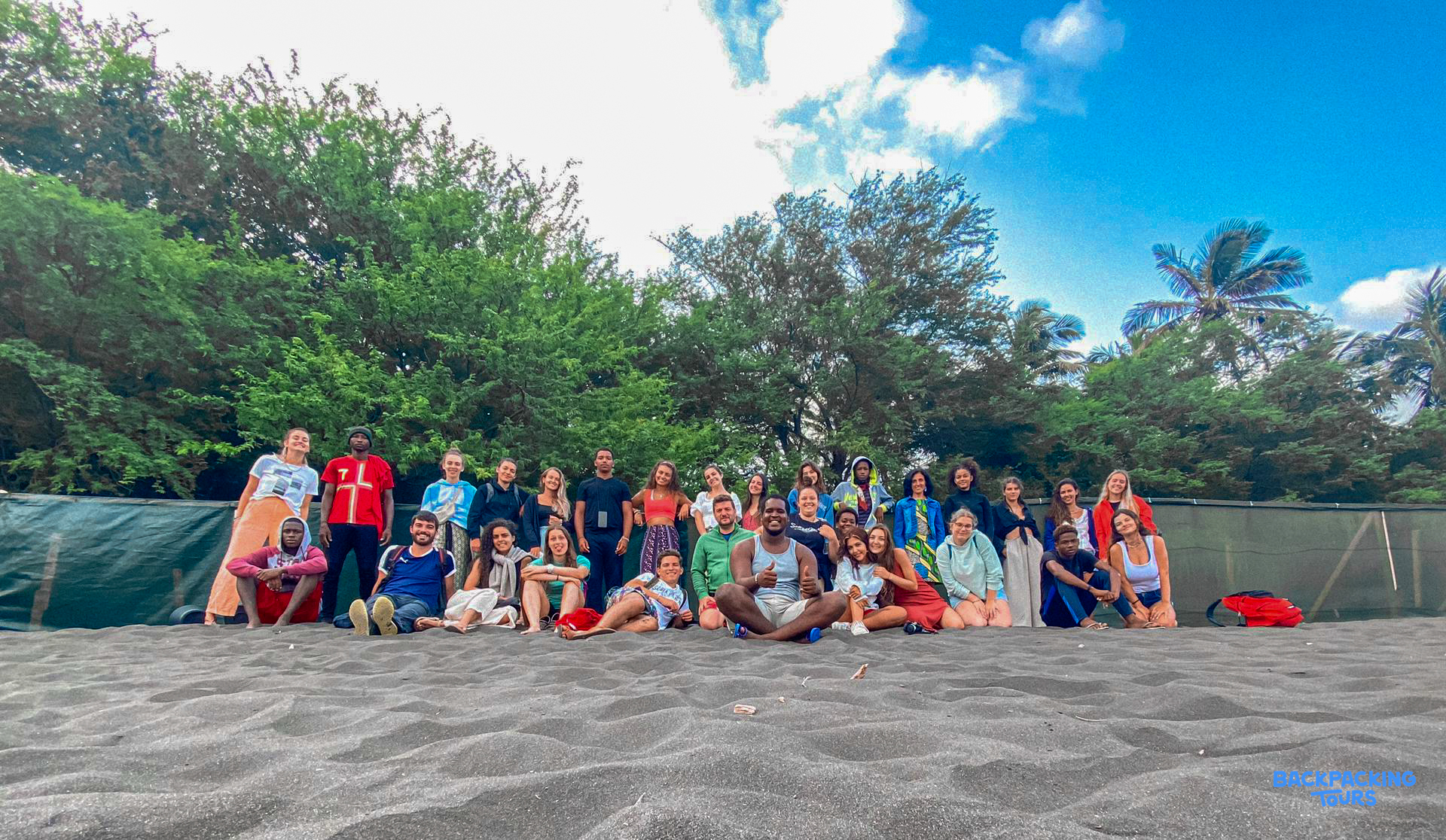 Happy travelers gather on a dark sandy beach, smiling for a group photo under a blue sky.