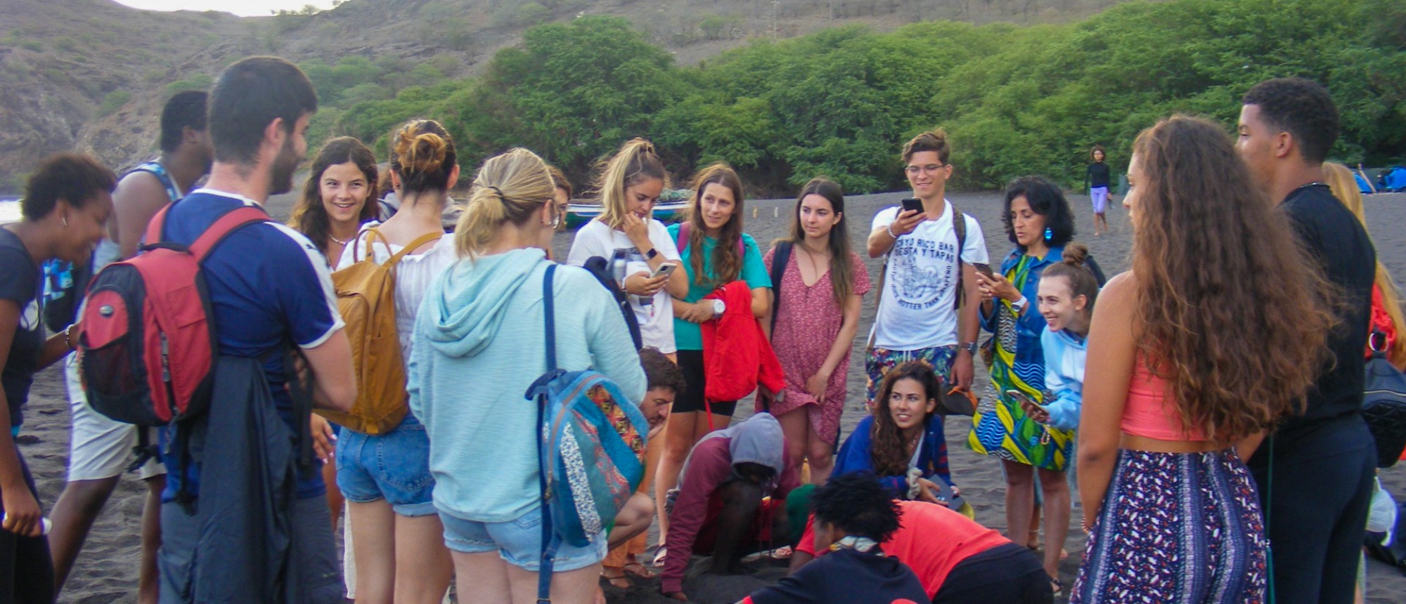 Young people and locals gather on a dark sand beach, observing a conservation activity.