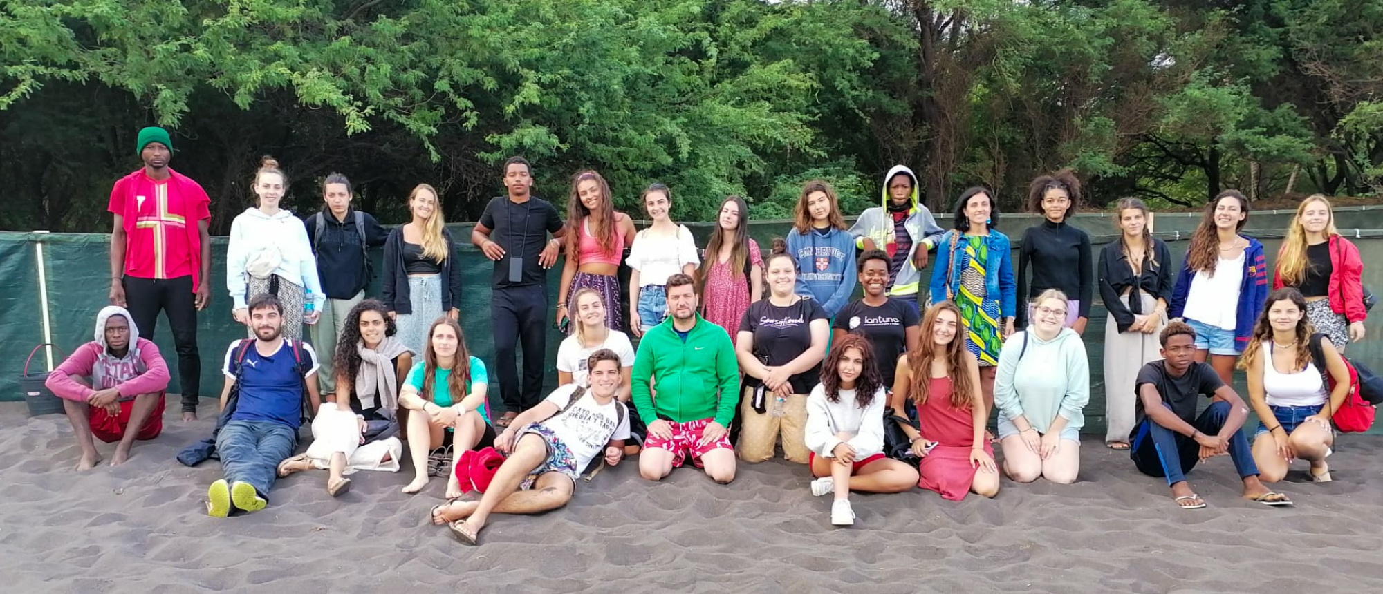A diverse group of young adults smiling for a group photo on dark sand with green trees.