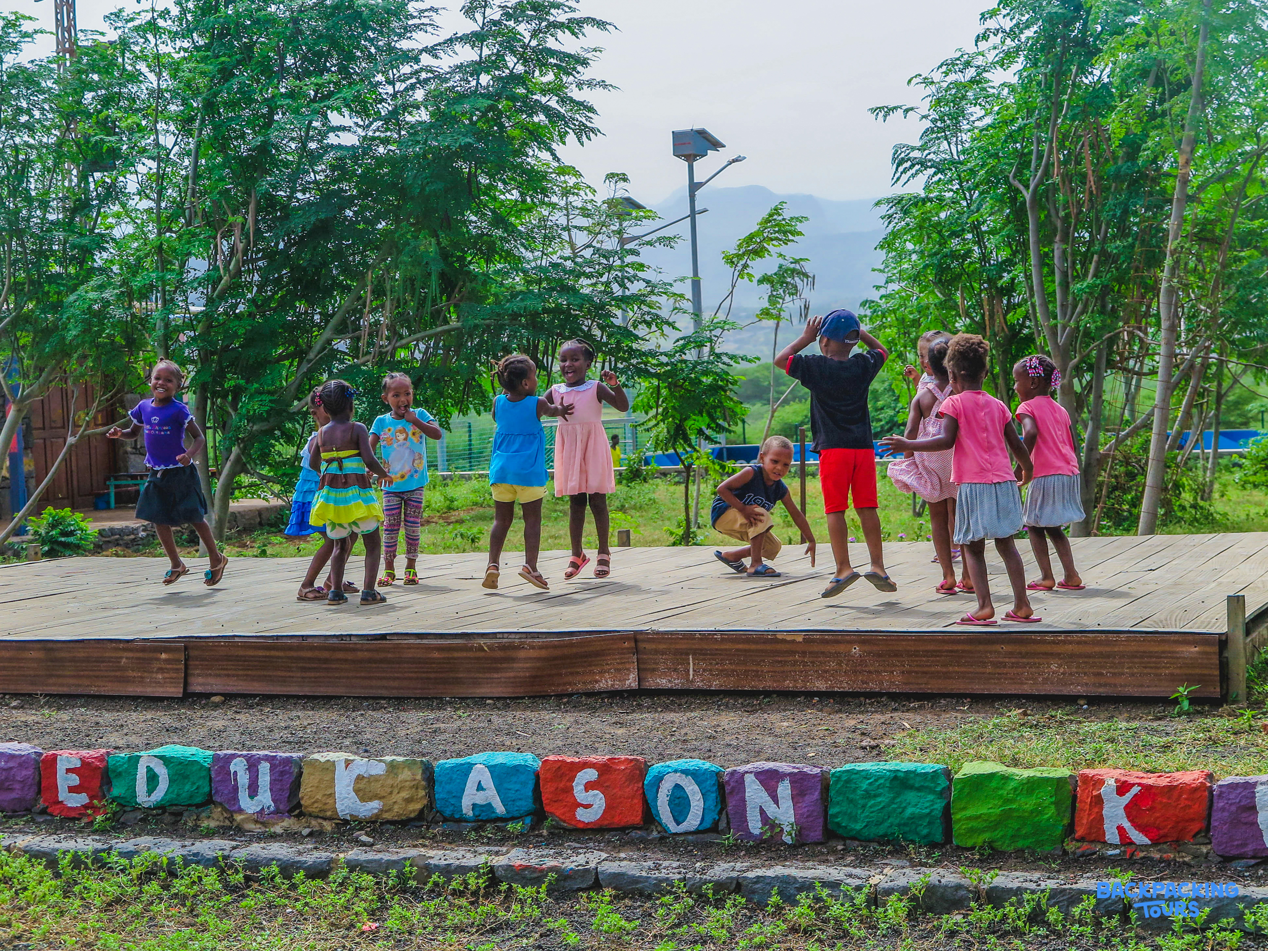 Young children joyfully jump and dance on an outdoor stage, with colorful rocks spelling 'EDUCASON K'.