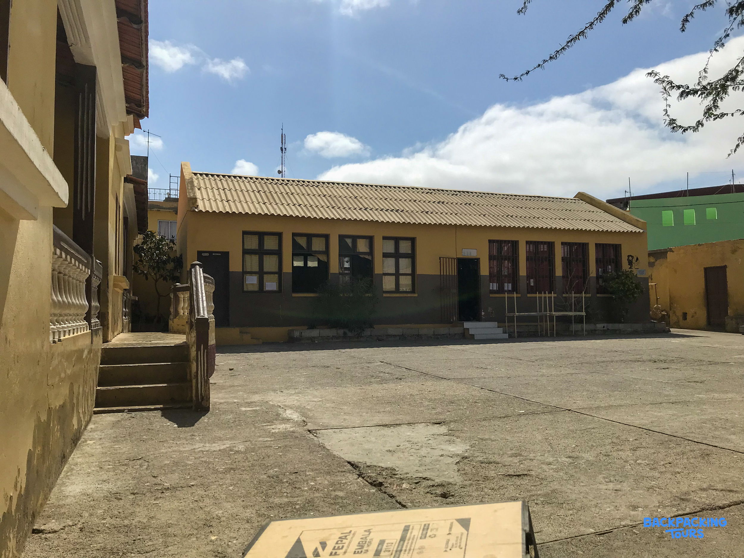 A yellow building with a tiled roof and many windows overlooks a large, empty paved courtyard.