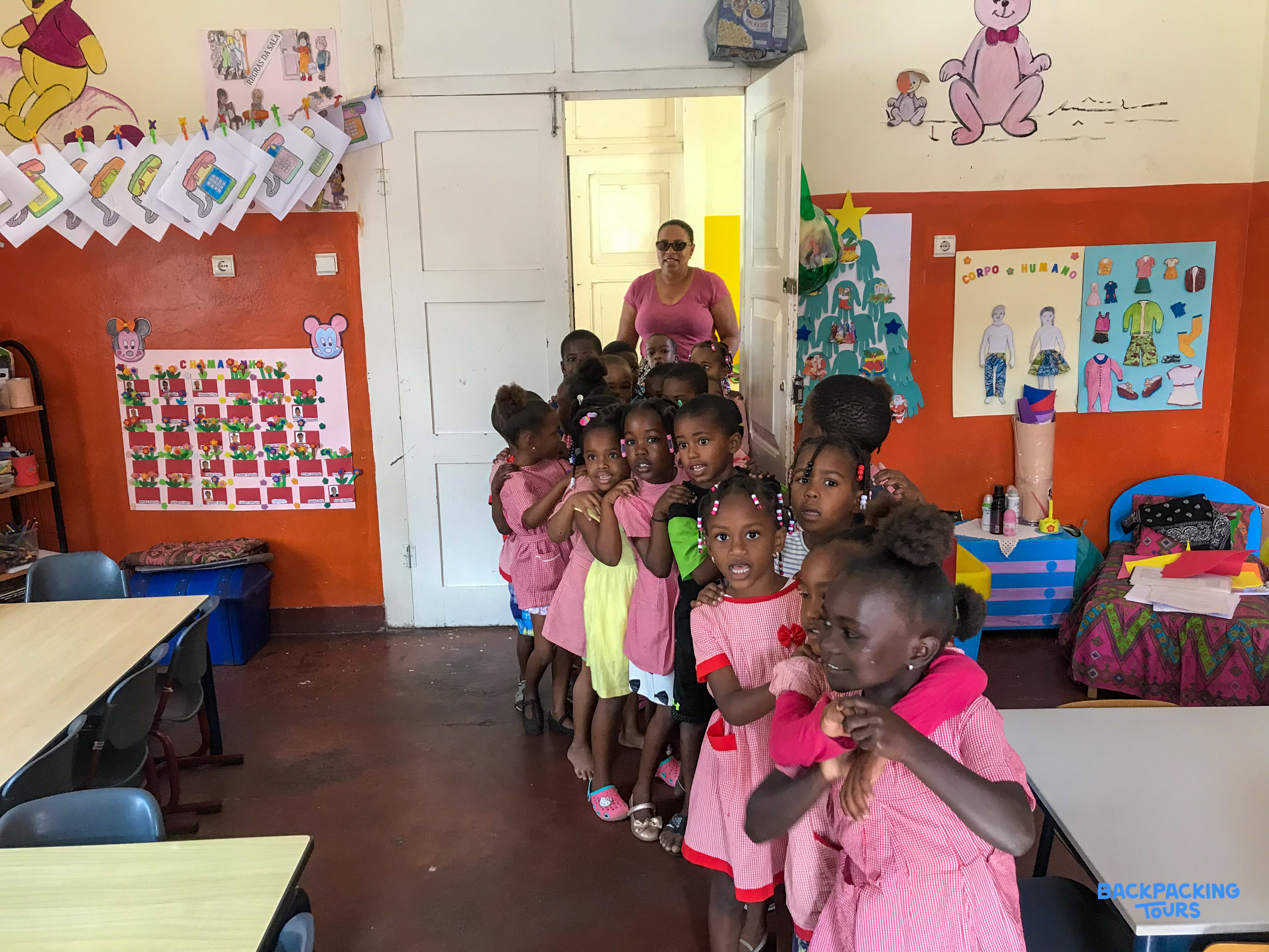 A line of smiling young children in pink dresses eagerly greet their teacher in a colorful classroom.