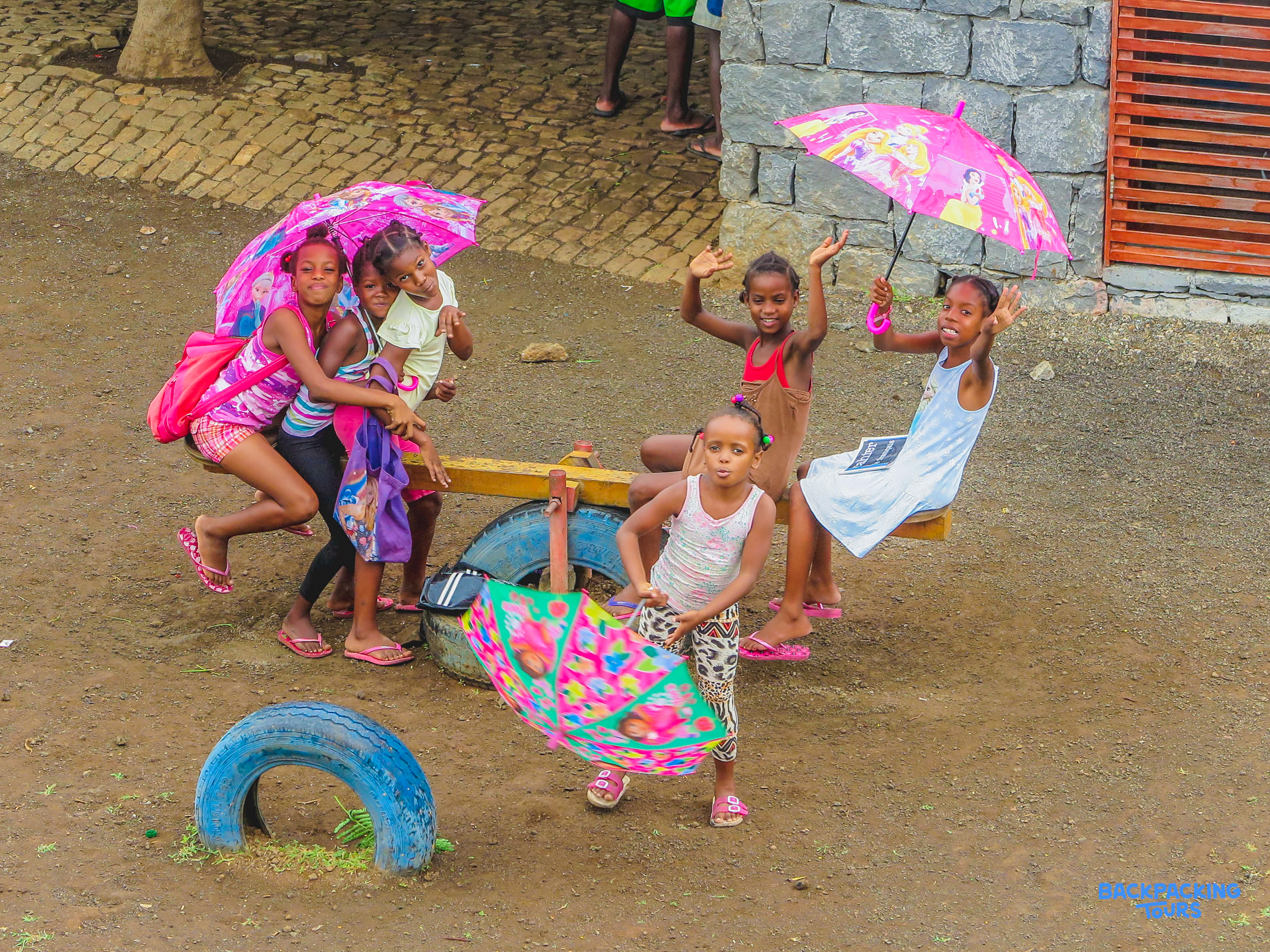 Smiling girls play with pink umbrellas and a makeshift seesaw in a vibrant outdoor scene.