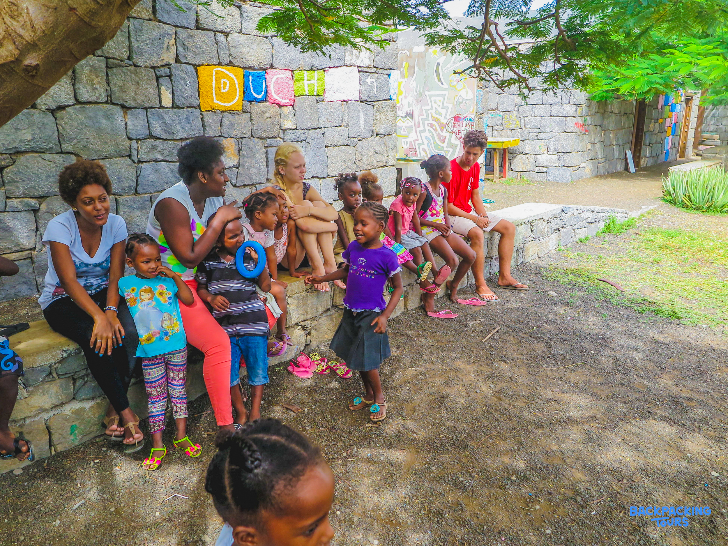 Adults and children relax outdoors under a tree, some braiding hair, in a warm Cape Verde community.