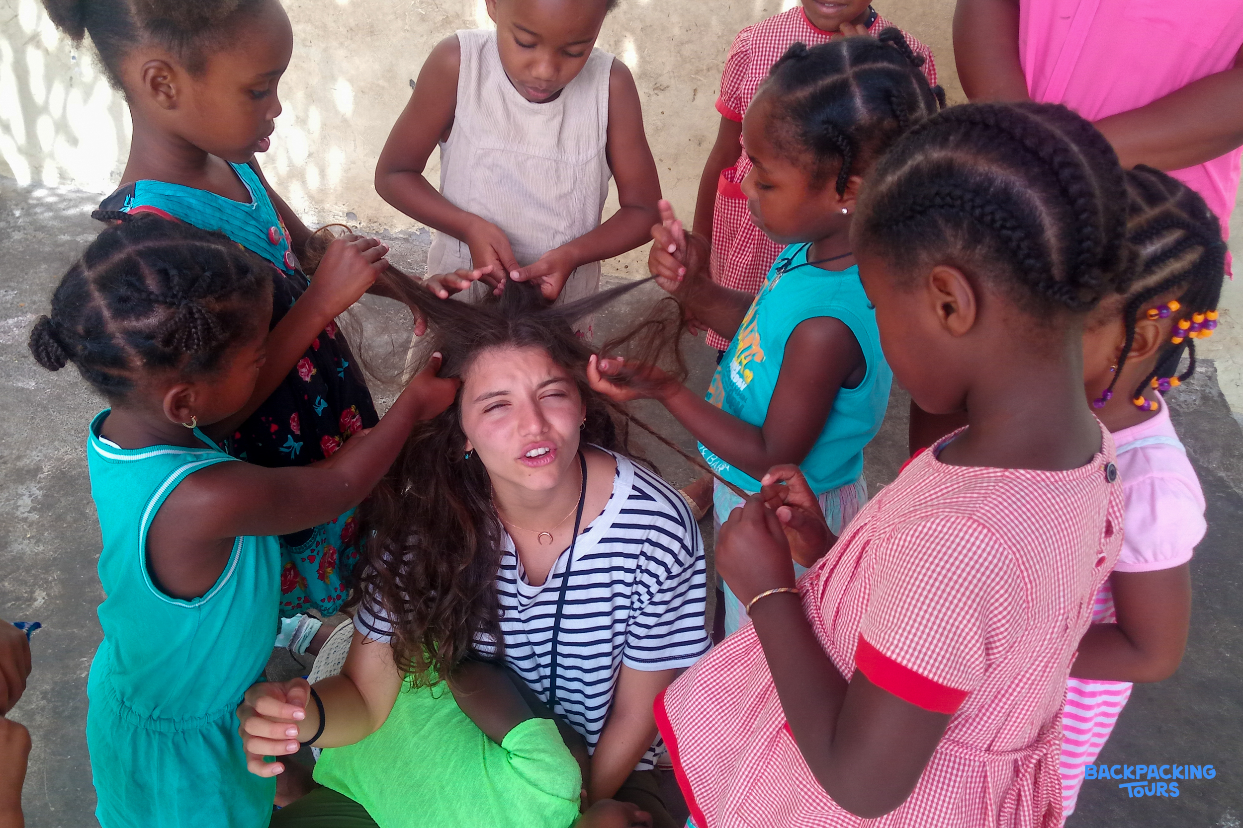 Young children playfully braid and style a volunteer's long curly hair outdoors, sharing a joyful moment.