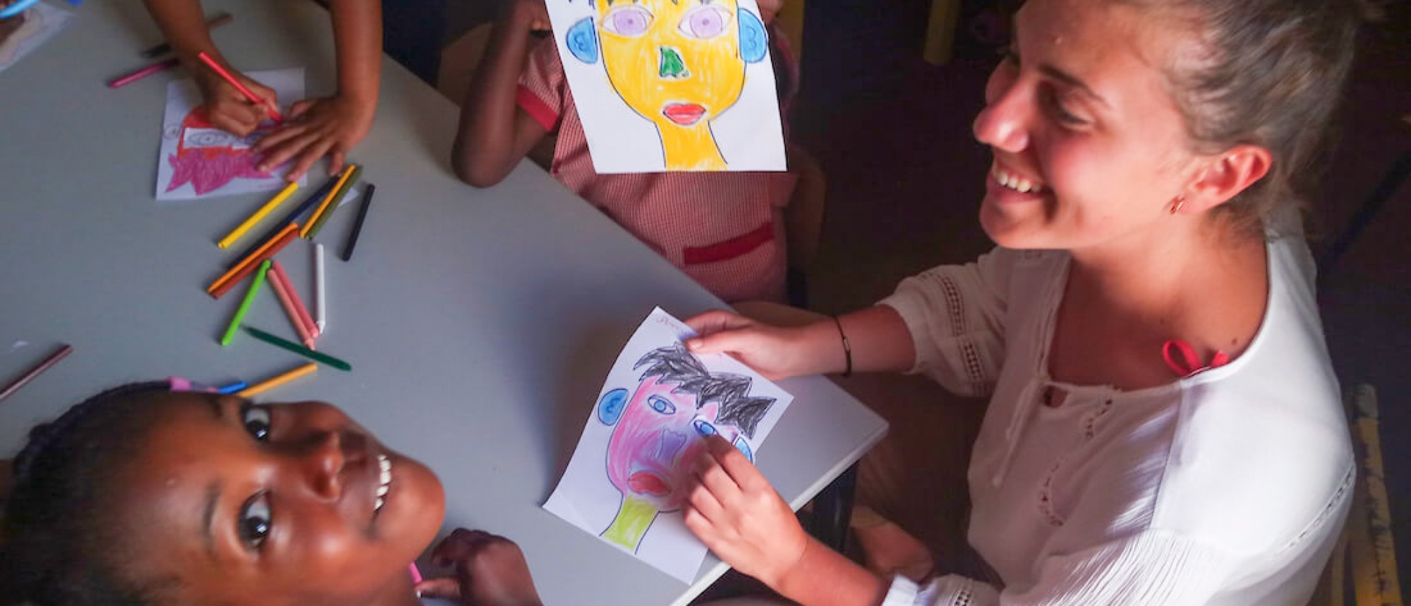 A smiling teacher and children coloring vibrant drawings together at a table.