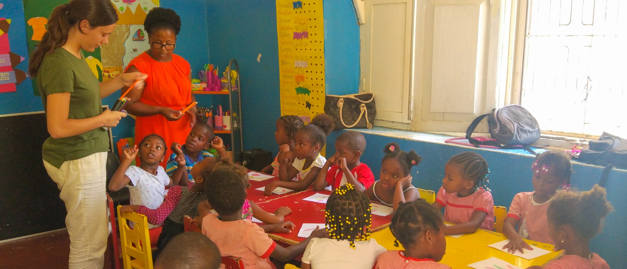 Two women teaching a group of young children seated at colorful tables in a bright classroom.