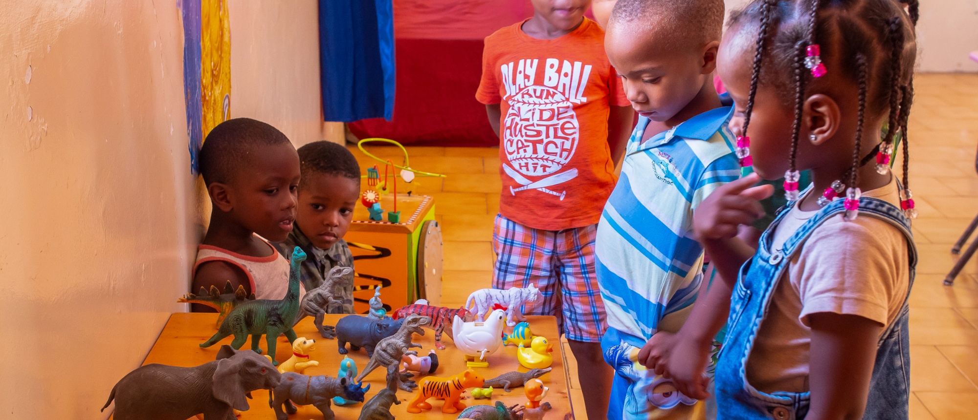 Curious kindergarten children gather around a table, exploring a variety of animal and dinosaur toys.