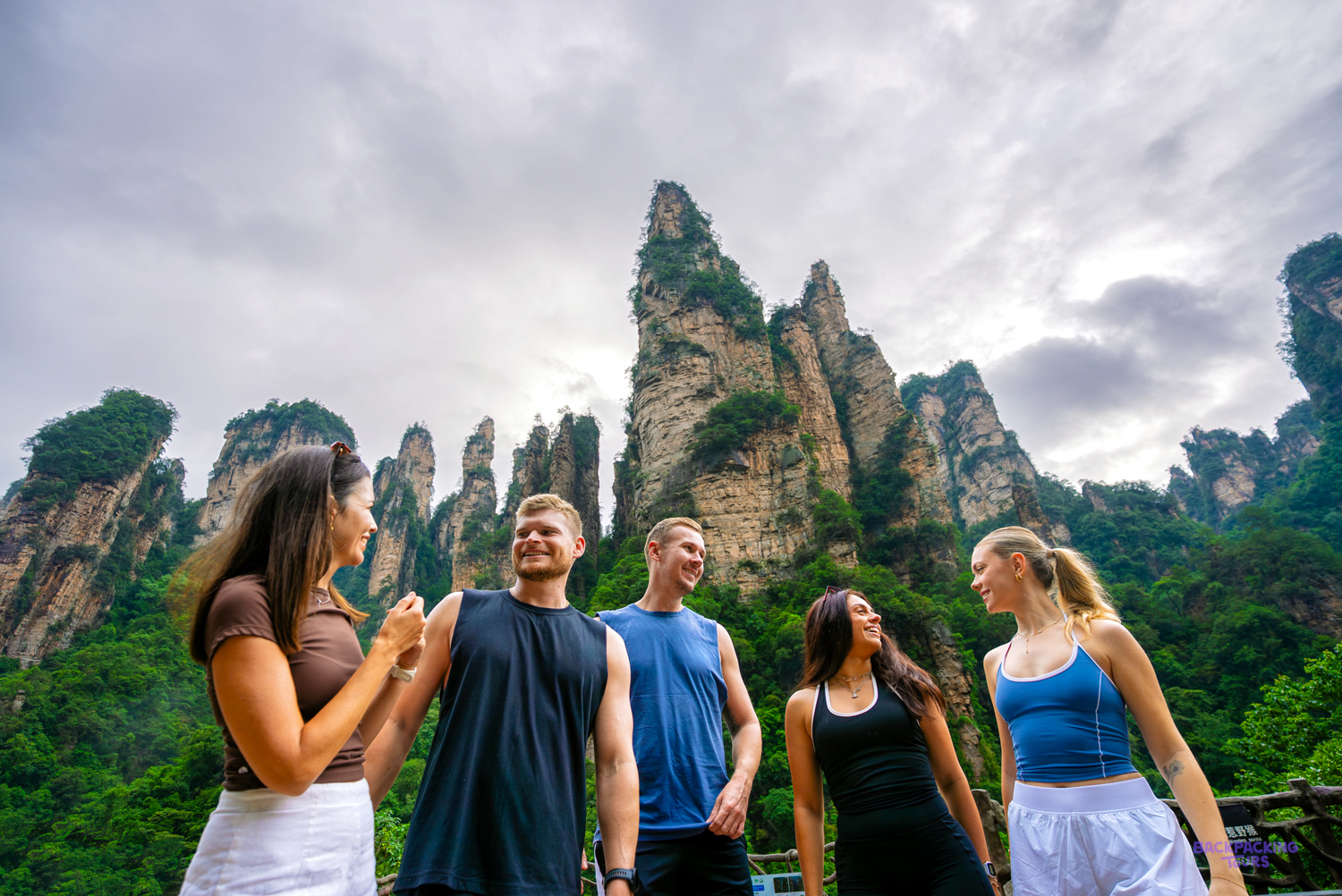 A group of travellers chats on a viewpoint in front of tall, tree-covered sandstone pillars.