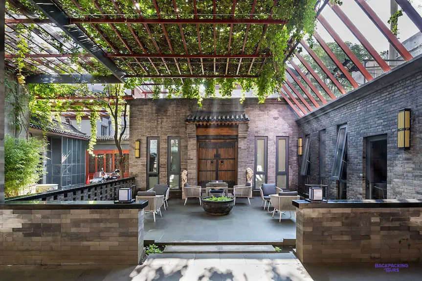 A hotel courtyard shaded by a vine-covered pergola, with seating and grey brick walls.