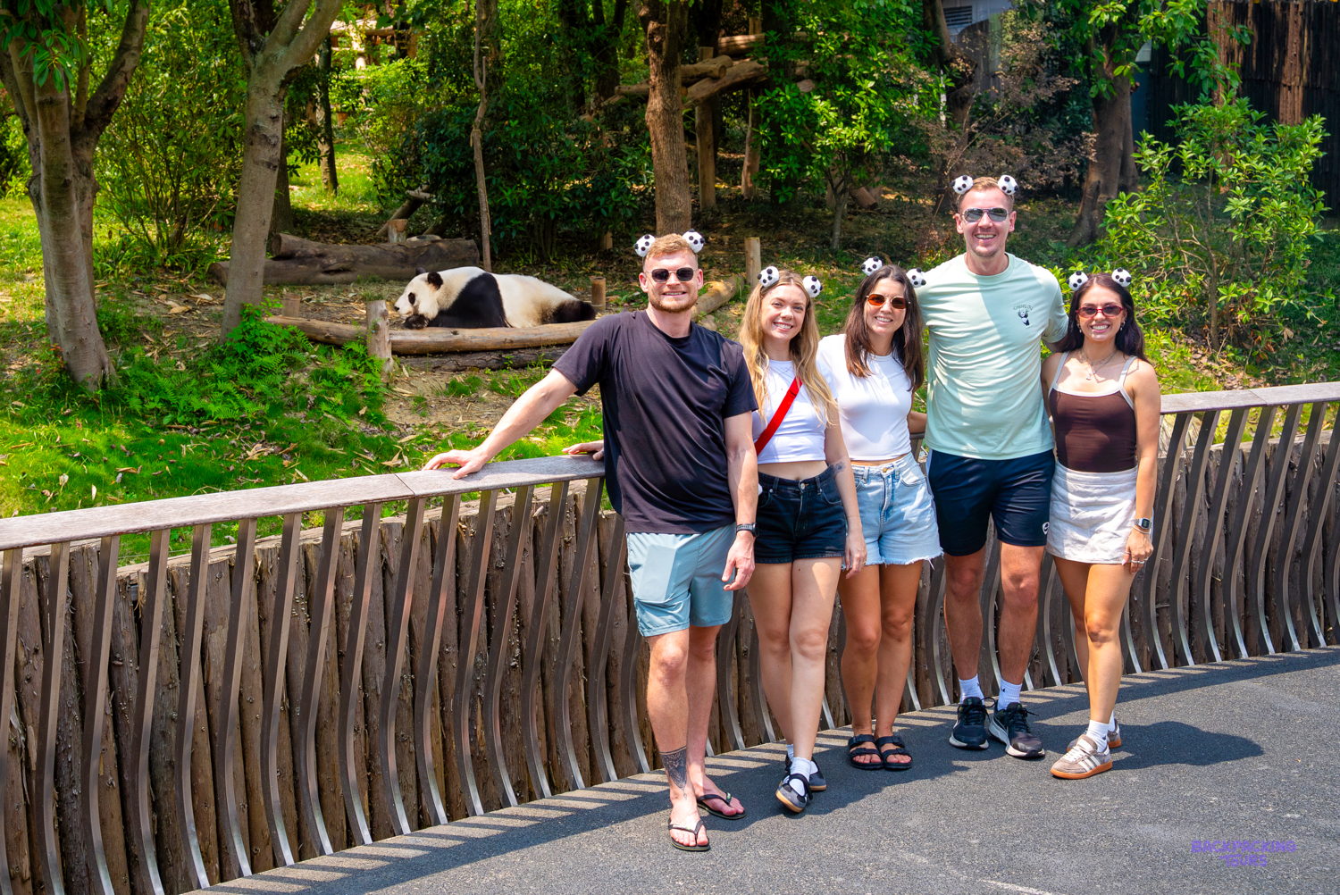 Five travellers pose by a wooden fence while a giant panda rests on a platform behind them.