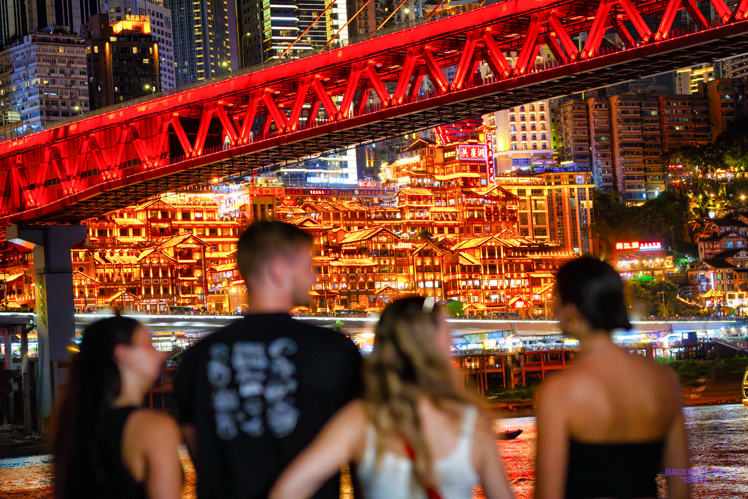 Travellers look across at a brightly lit cliffside building beneath a red steel bridge at night.
