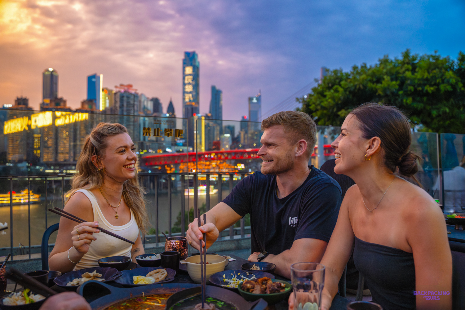 Three travellers share a hotpot at an outdoor table as the city skyline lights up at sunset.