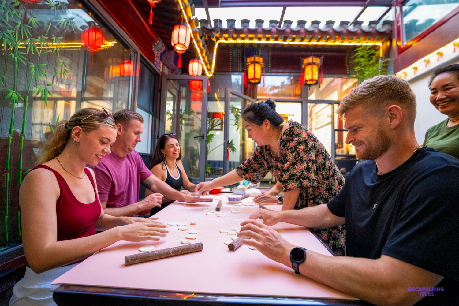 Travellers fold dumplings around a long table with a local teacher, beneath red paper lanterns.