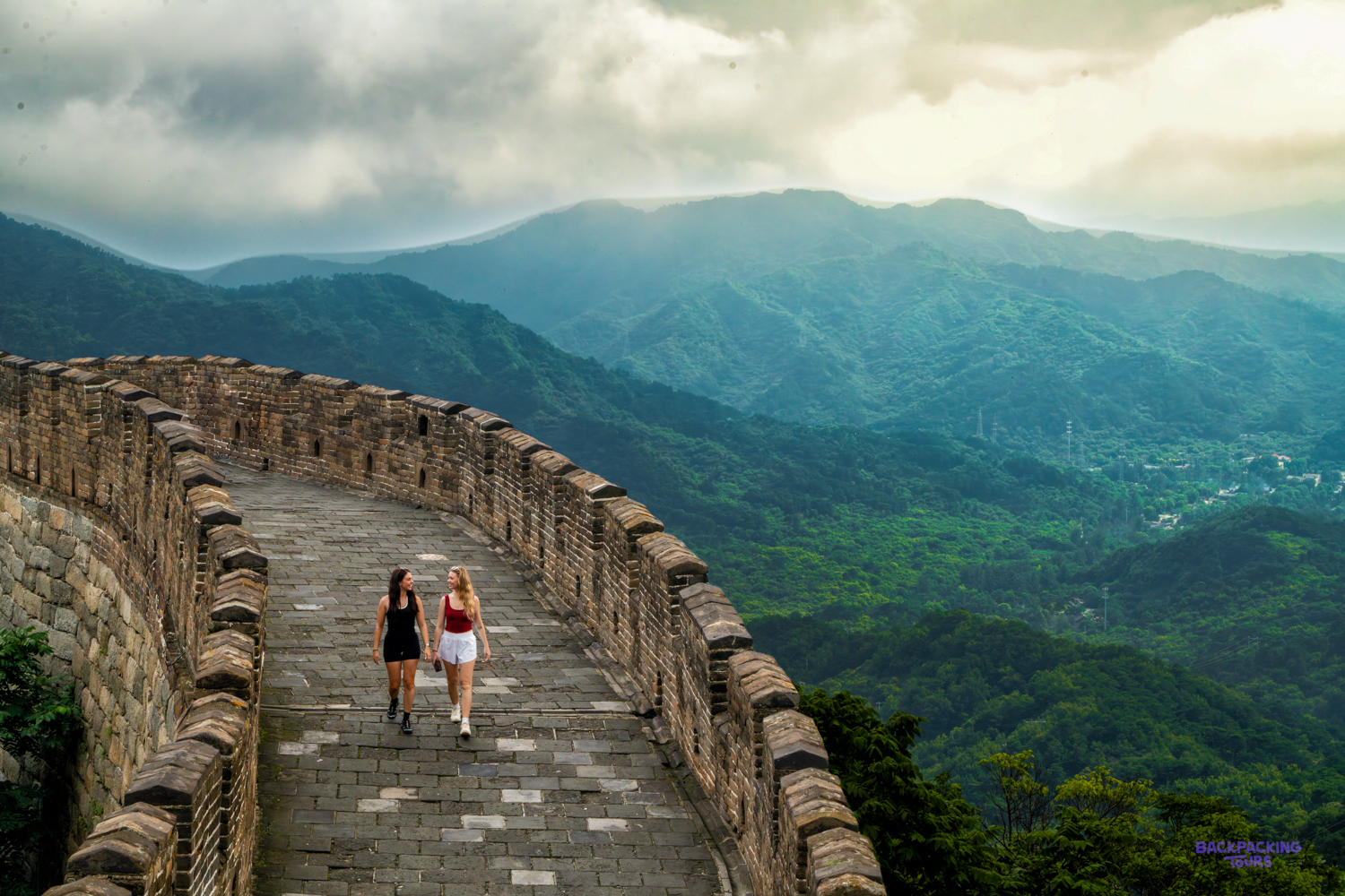 Two travellers walk along the Great Wall as it follows a forested ridge into misty mountains.