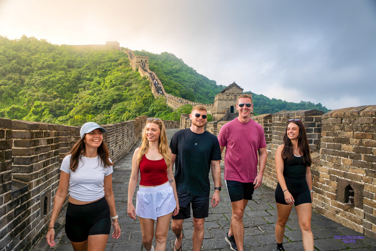 A group of travellers walks along the Great Wall with green hills and a watchtower behind them.