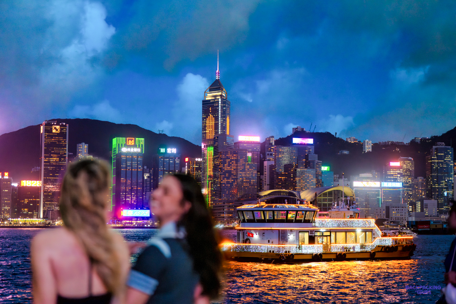 Two travellers watch a lit-up ferry cross the harbour below Hong Kong's glowing skyscrapers at dusk.
