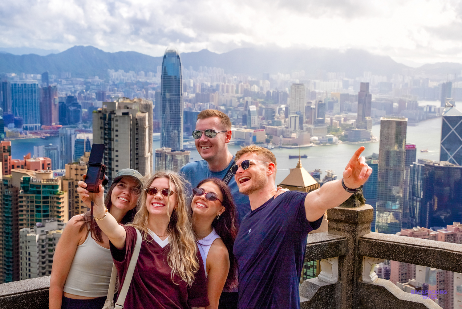 A group of travellers takes a selfie on a viewpoint high above Hong Kong's skyscrapers and harbour.