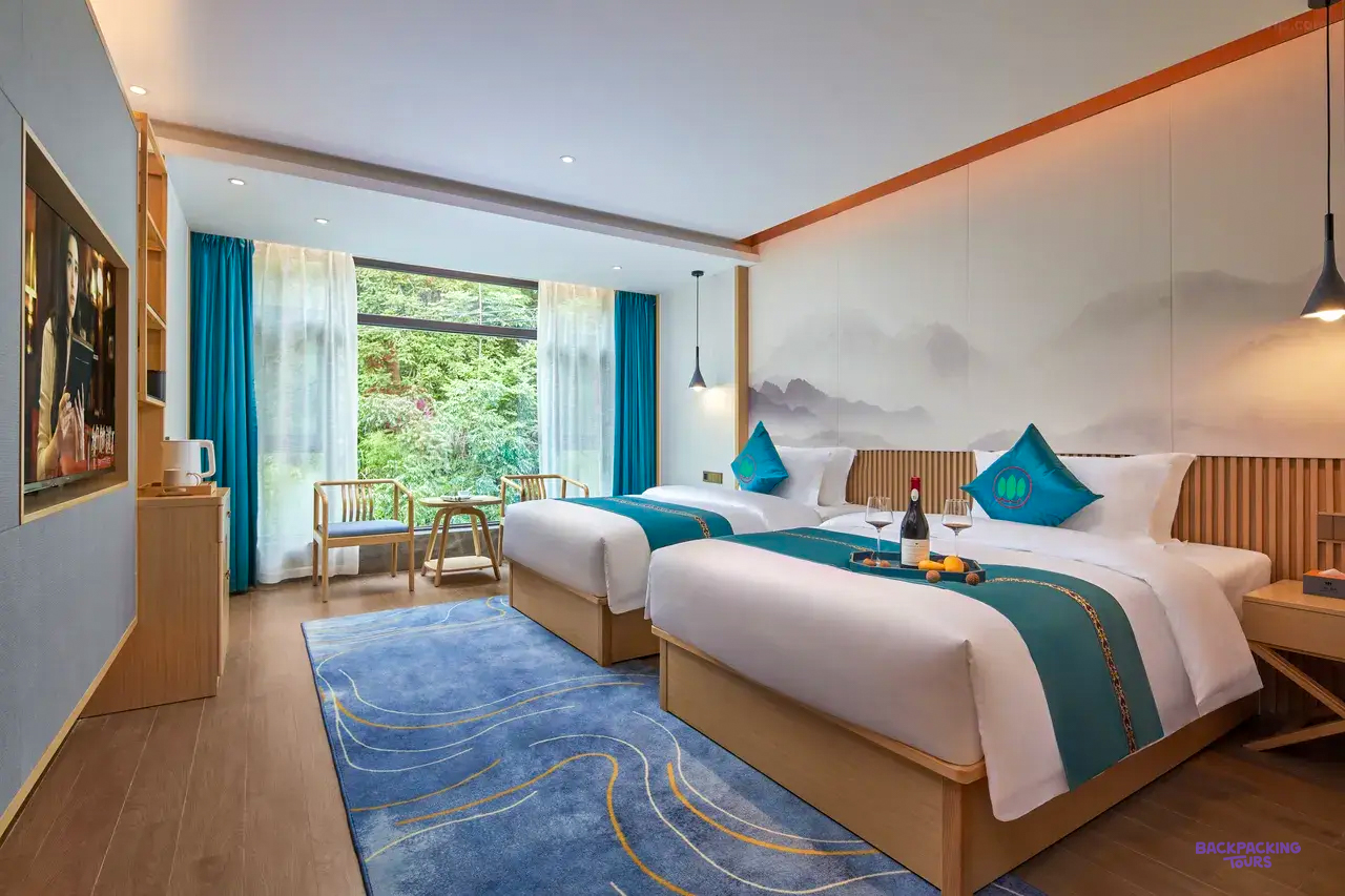 A twin room with teal cushions, a blue rug and a large window onto green trees.