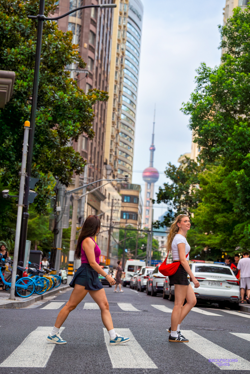 Two travellers cross a tree-lined street in Shanghai with the Oriental Pearl Tower in the distance.