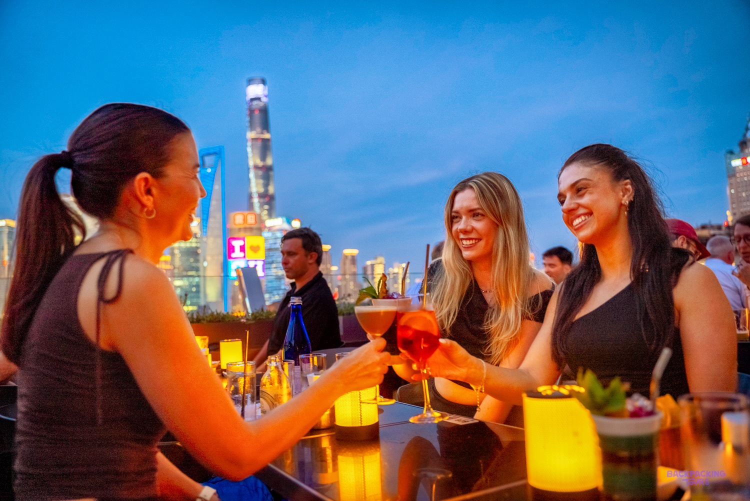 Three friends raise colourful cocktails at a rooftop bar with the Shanghai skyline behind them at dusk.