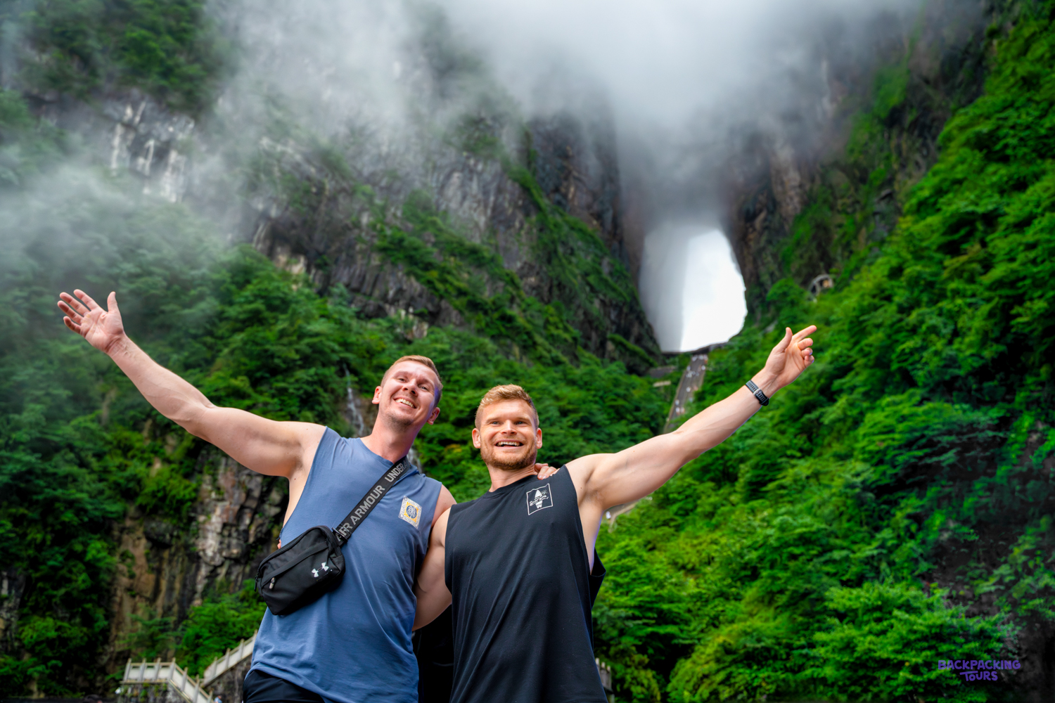 Two travellers throw their arms wide beneath a huge natural arch in a green, misty mountainside.