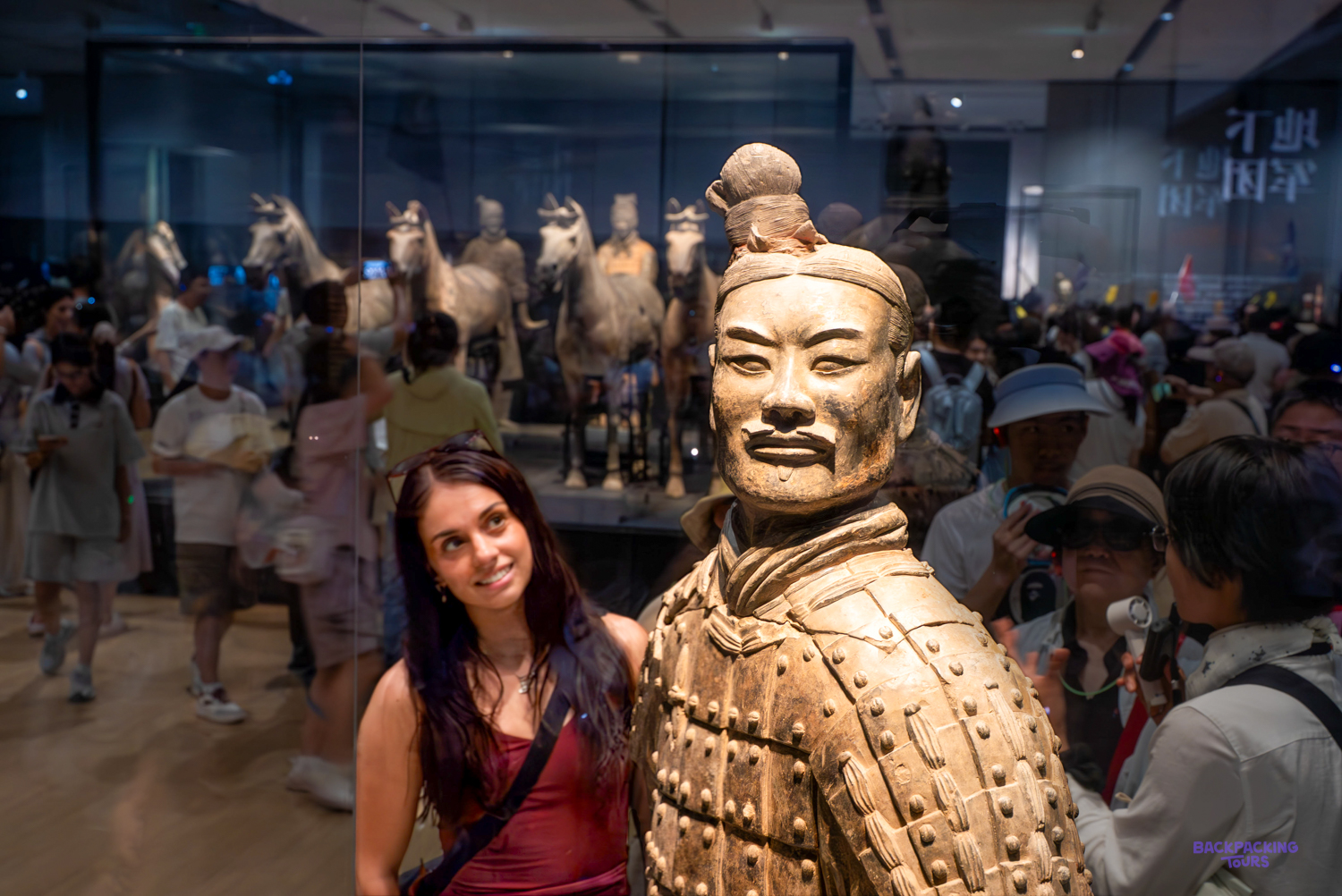 A traveller smiles beside a terracotta warrior figure in a busy museum hall, with more figures behind.