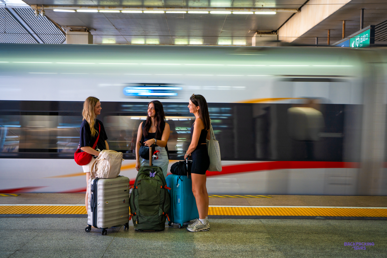 Three travellers with backpacks and suitcases wait on a platform as a high-speed train speeds past.