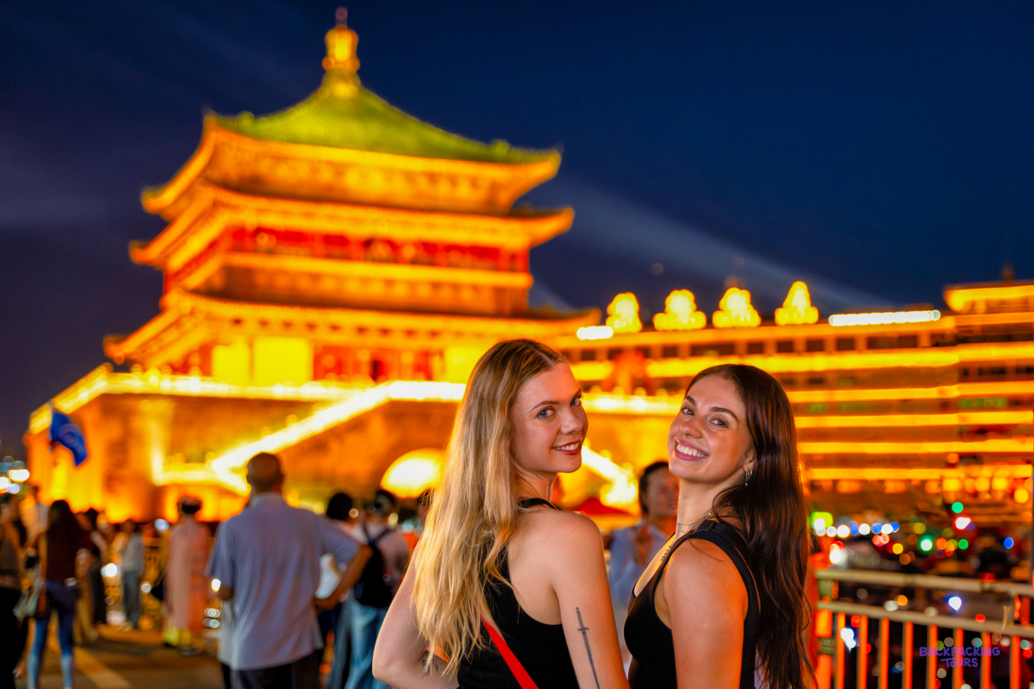 Two travellers smile in front of a brightly lit traditional tower on a busy evening street.