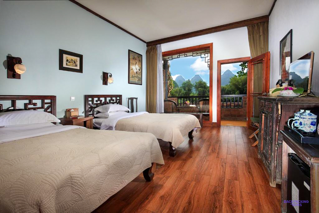 A twin room with carved wooden beds and an open balcony door looking out to limestone peaks.