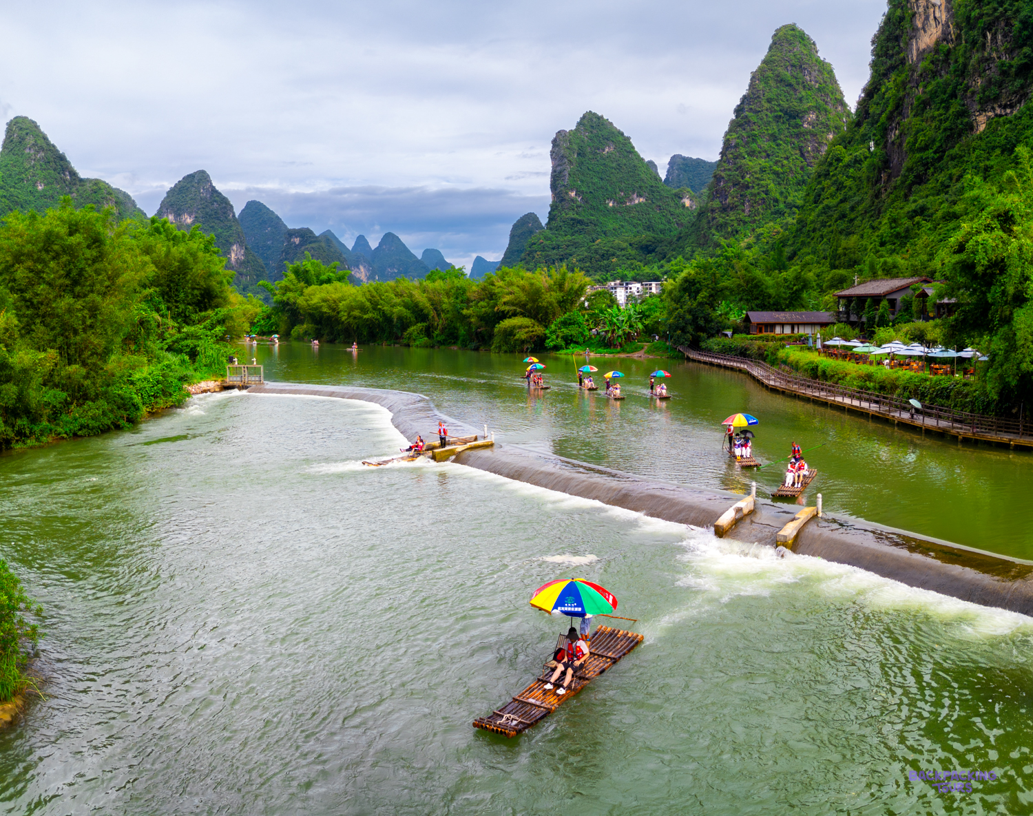Bamboo rafts drift down a green river between limestone peaks, seen from above.