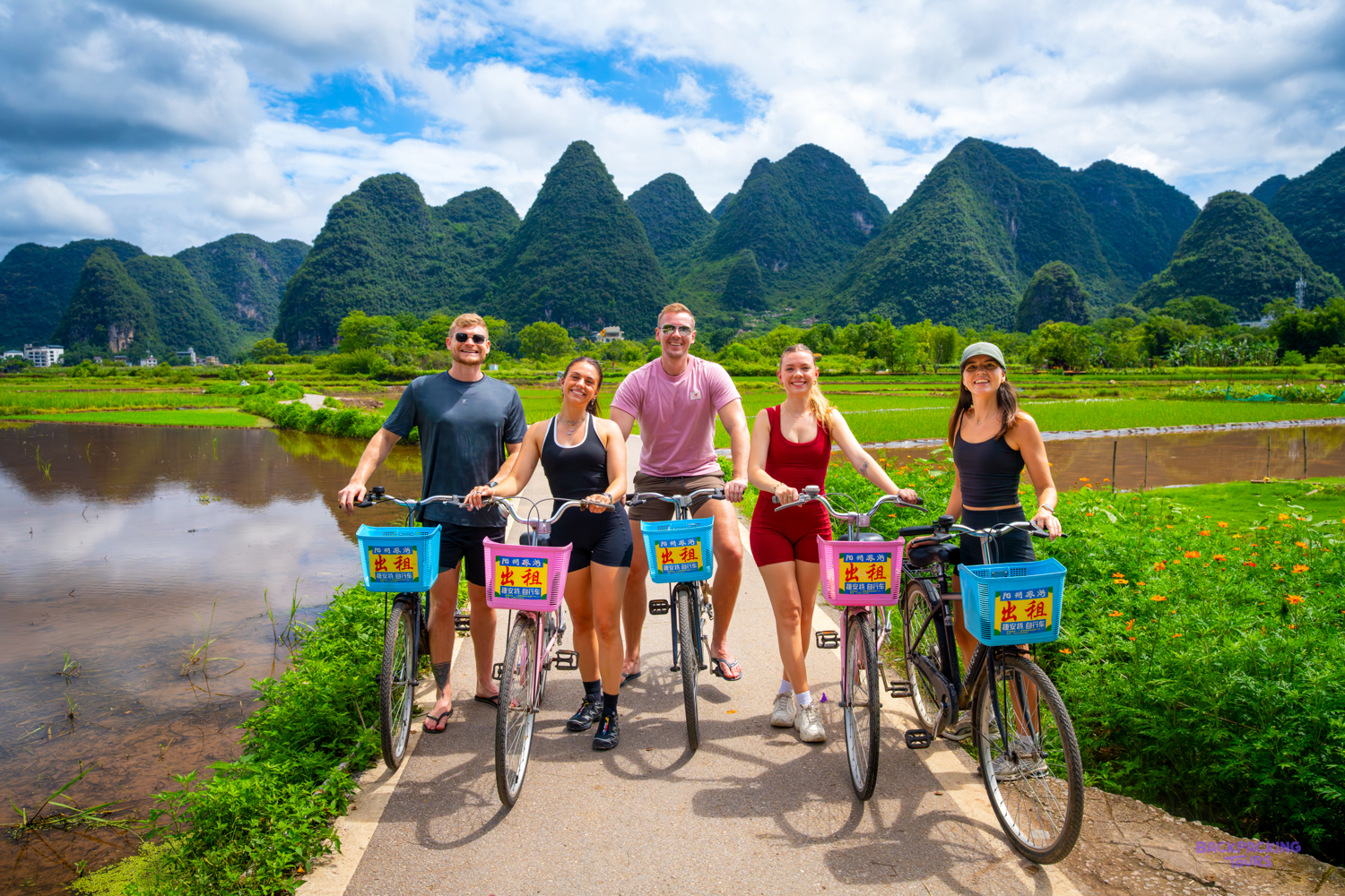 A group of travellers stands with bicycles on a country path beside rice paddies and limestone peaks.