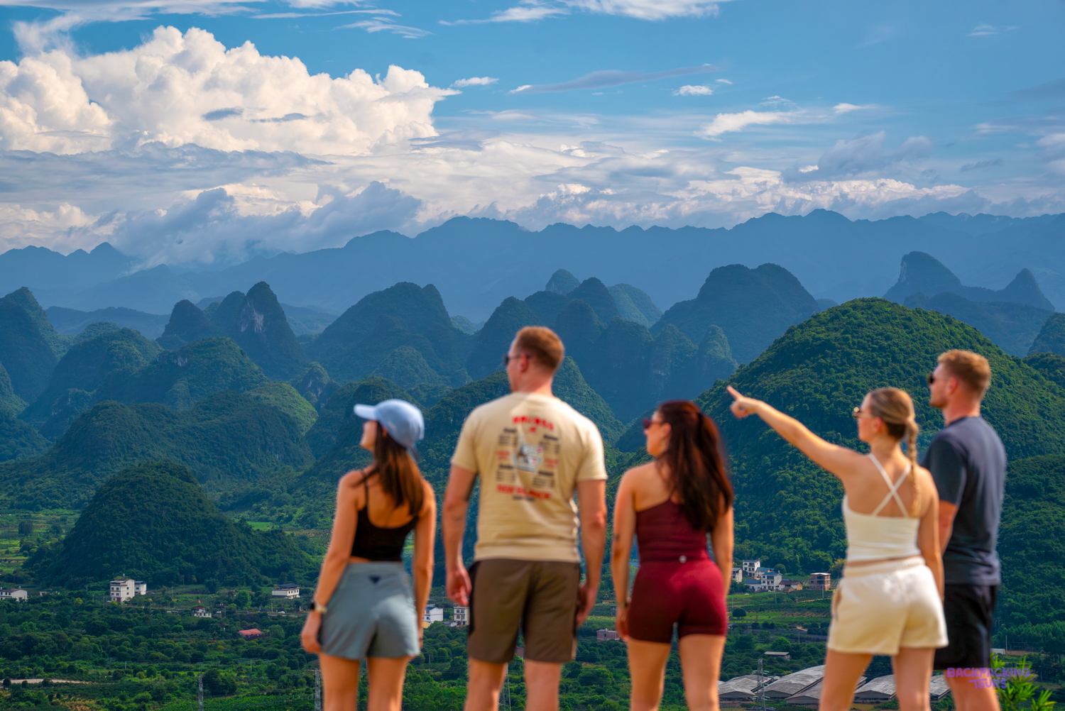 Travellers look out from a hilltop over a sea of jagged green limestone peaks under a blue sky.