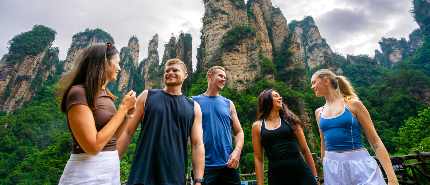 A group of travellers chats on a viewpoint in front of tall, tree-covered sandstone pillars.