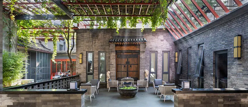 A hotel courtyard shaded by a vine-covered pergola, with seating and grey brick walls.