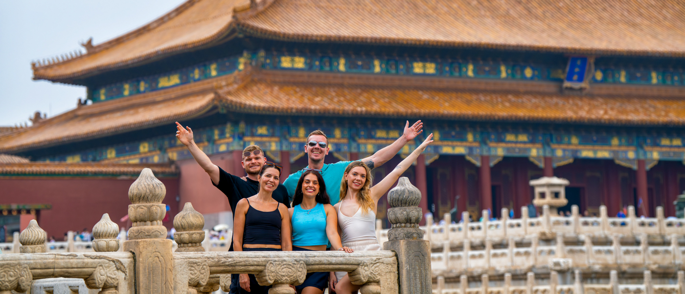 A group of travellers waves and cheers on a marble terrace in front of a golden-roofed palace hall.