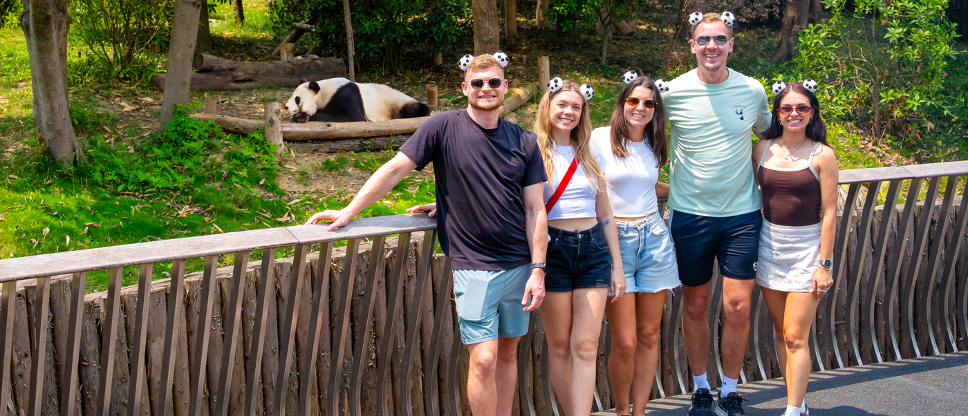 Five travellers pose by a wooden fence while a giant panda rests on a platform behind them.