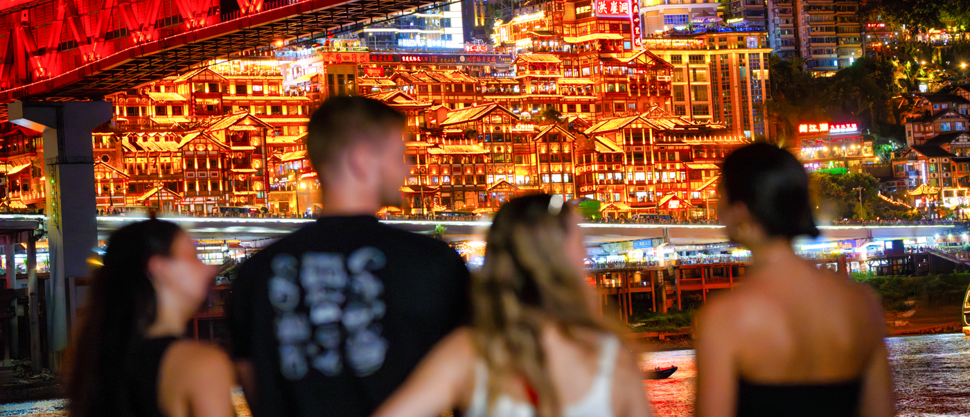 Travellers look across at a brightly lit cliffside building beneath a red steel bridge at night.