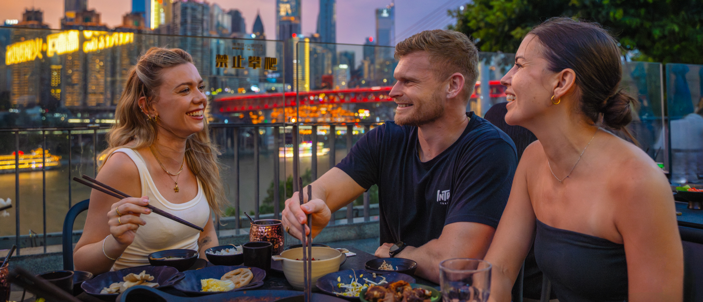 Three travellers share a hotpot at an outdoor table as the city skyline lights up at sunset.