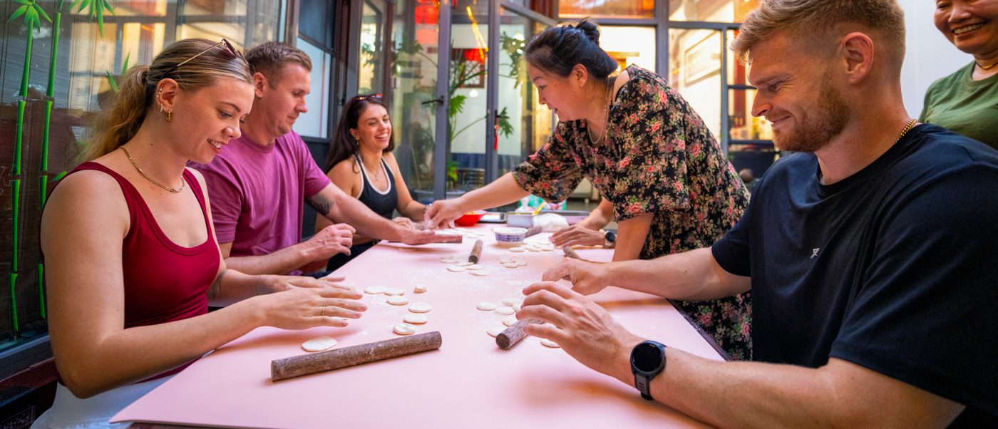 Travellers fold dumplings around a long table with a local teacher, beneath red paper lanterns.