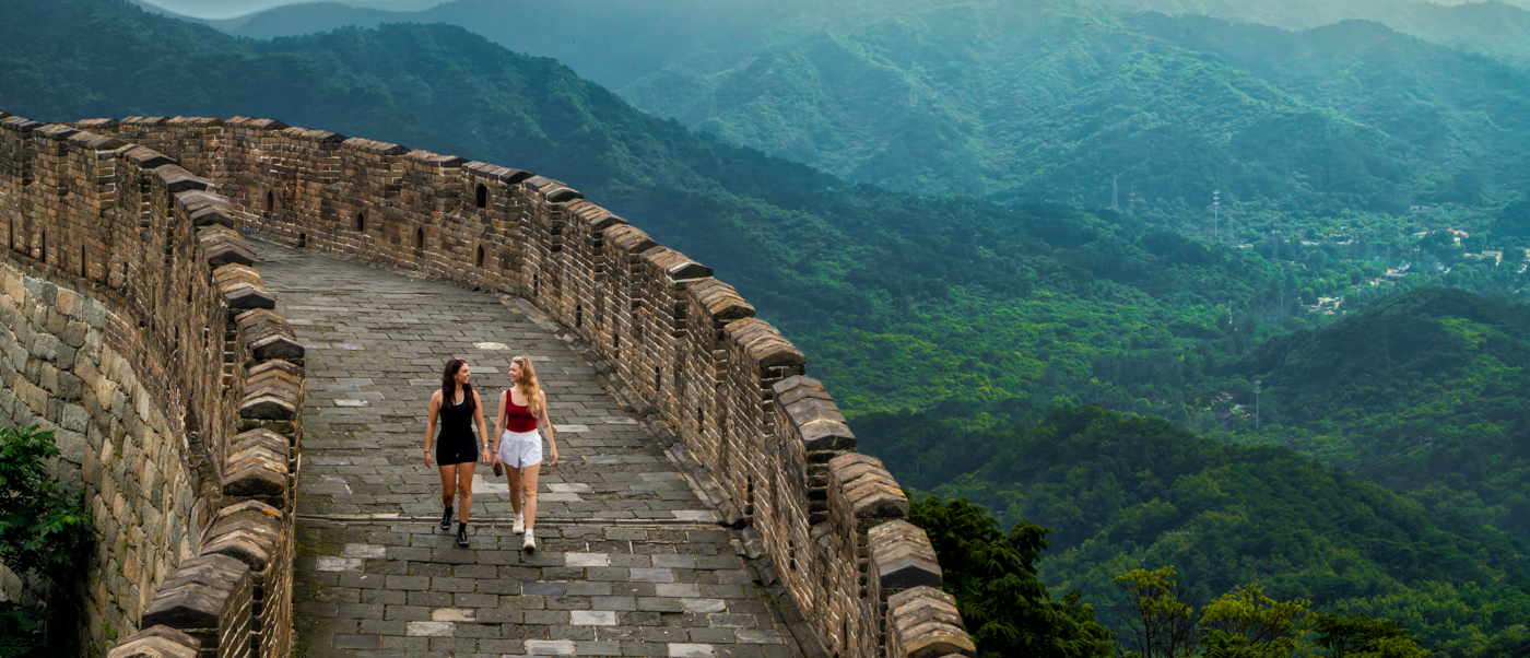 Two travellers walk along the Great Wall as it follows a forested ridge into misty mountains.