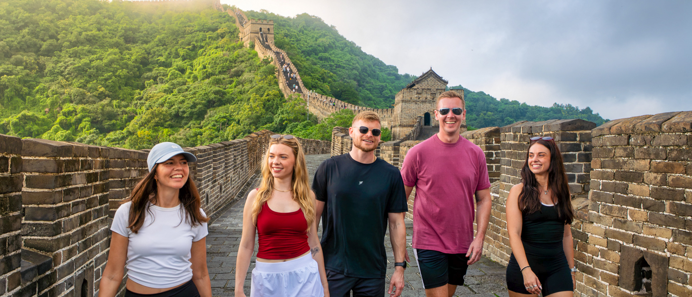 A group of travellers walks along the Great Wall with green hills and a watchtower behind them.
