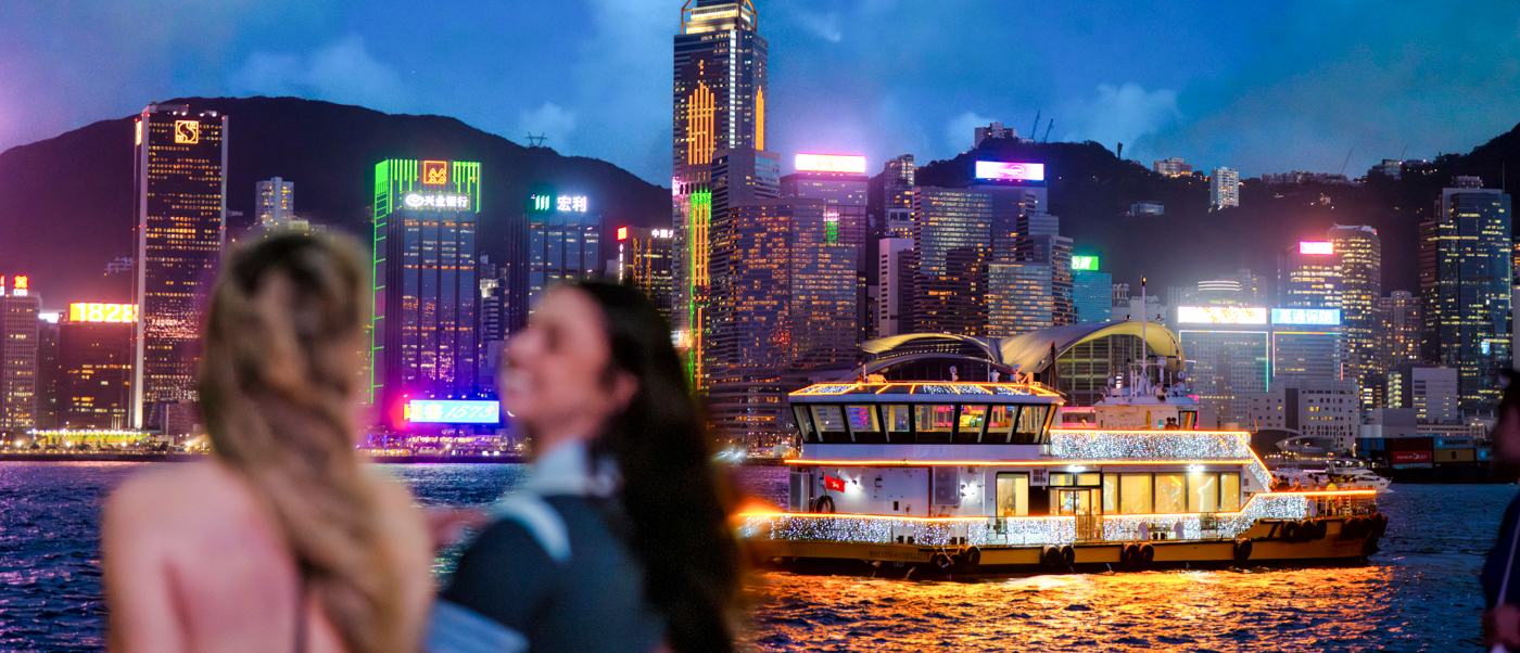 Two travellers watch a lit-up ferry cross the harbour below Hong Kong's glowing skyscrapers at dusk.