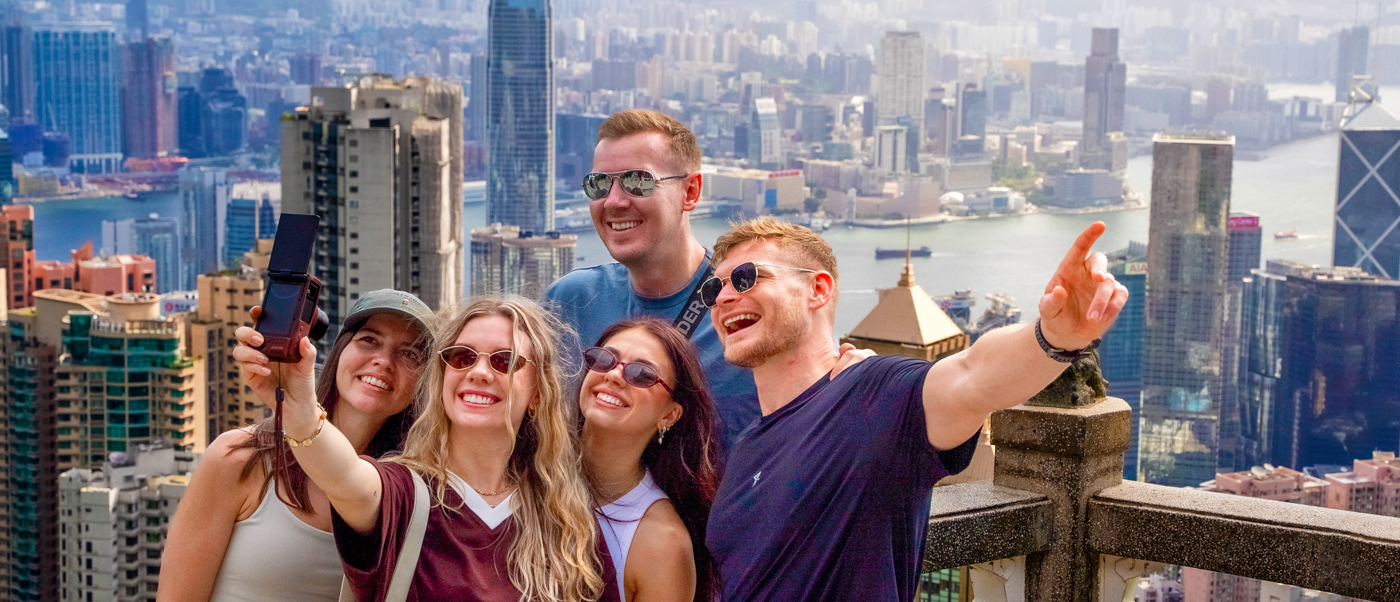 A group of travellers takes a selfie on a viewpoint high above Hong Kong's skyscrapers and harbour.