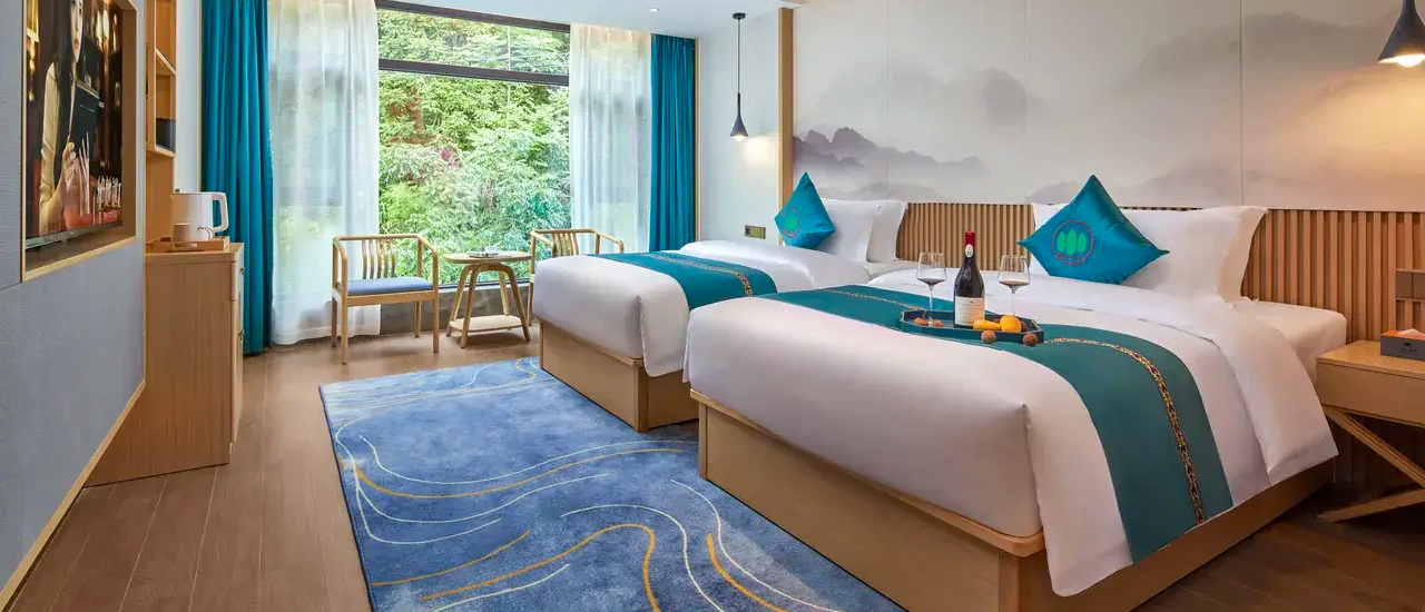 A twin room with teal cushions, a blue rug and a large window onto green trees.
