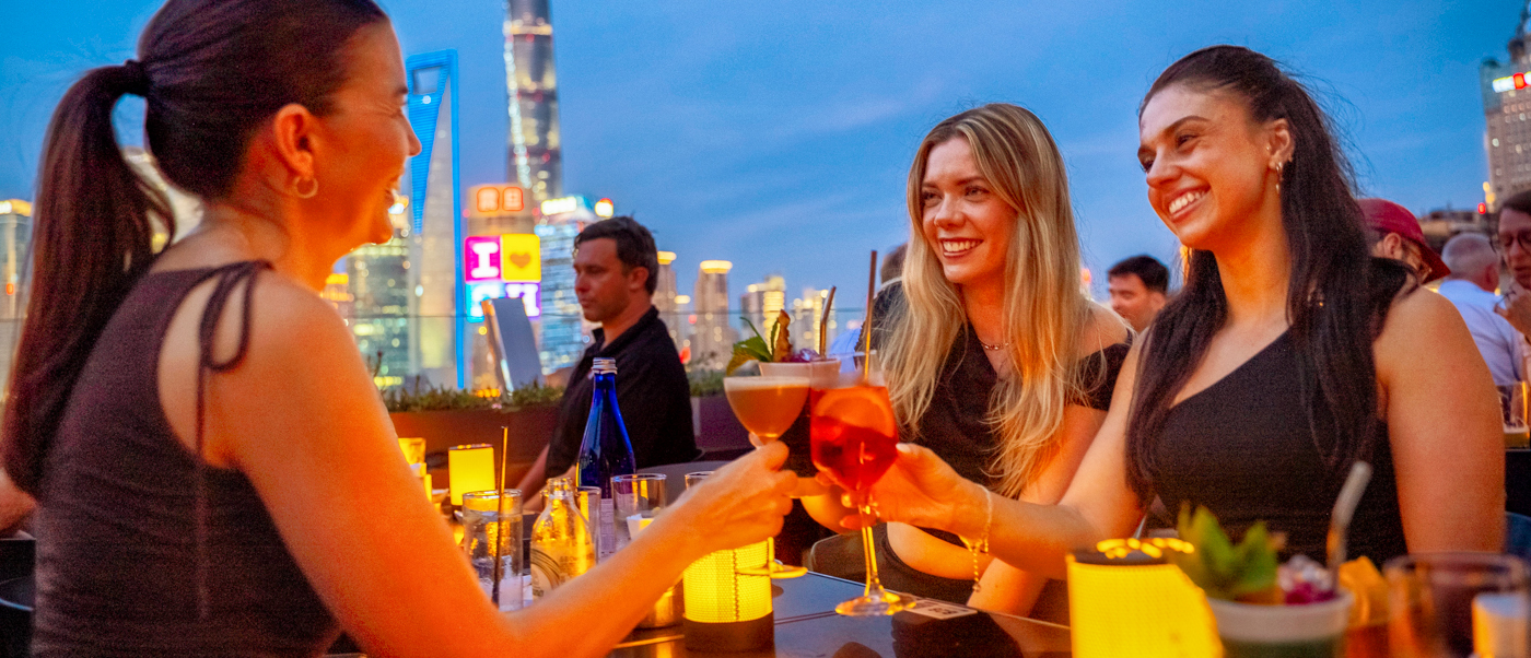 Three friends raise colourful cocktails at a rooftop bar with the Shanghai skyline behind them at dusk.
