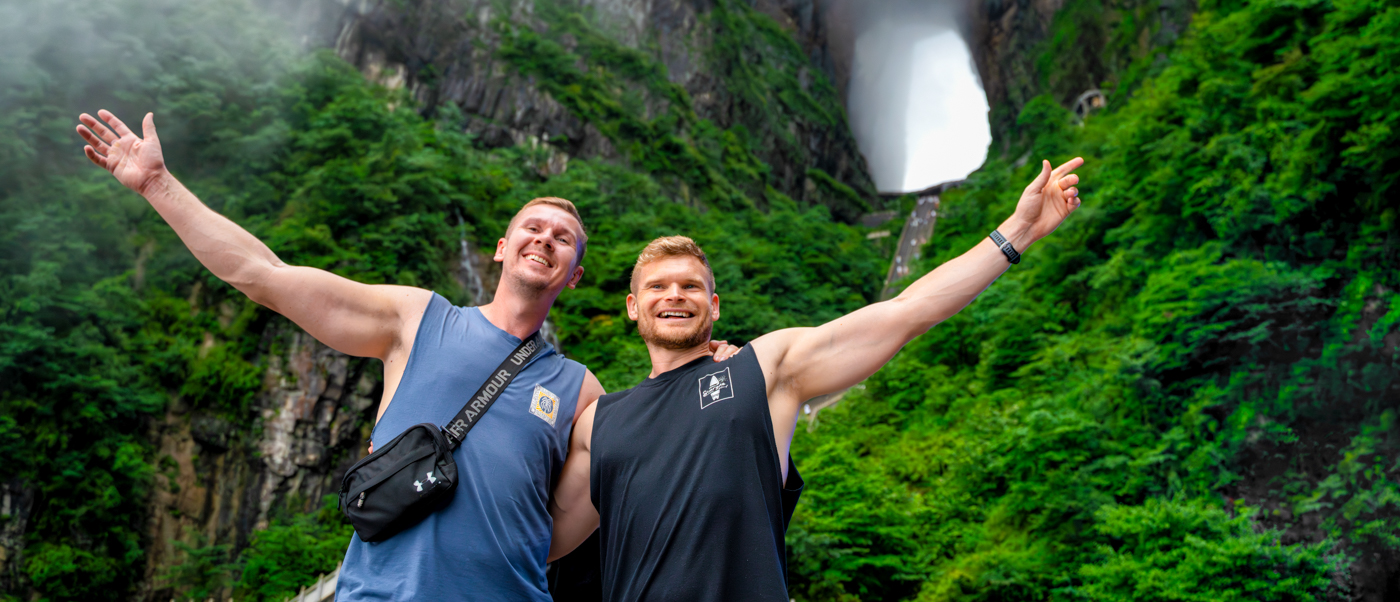 Two travellers throw their arms wide beneath a huge natural arch in a green, misty mountainside.