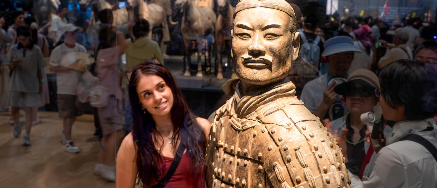 A traveller smiles beside a terracotta warrior figure in a busy museum hall, with more figures behind.