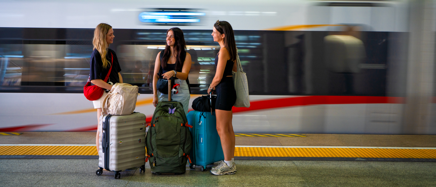 Three travellers with backpacks and suitcases wait on a platform as a high-speed train speeds past.