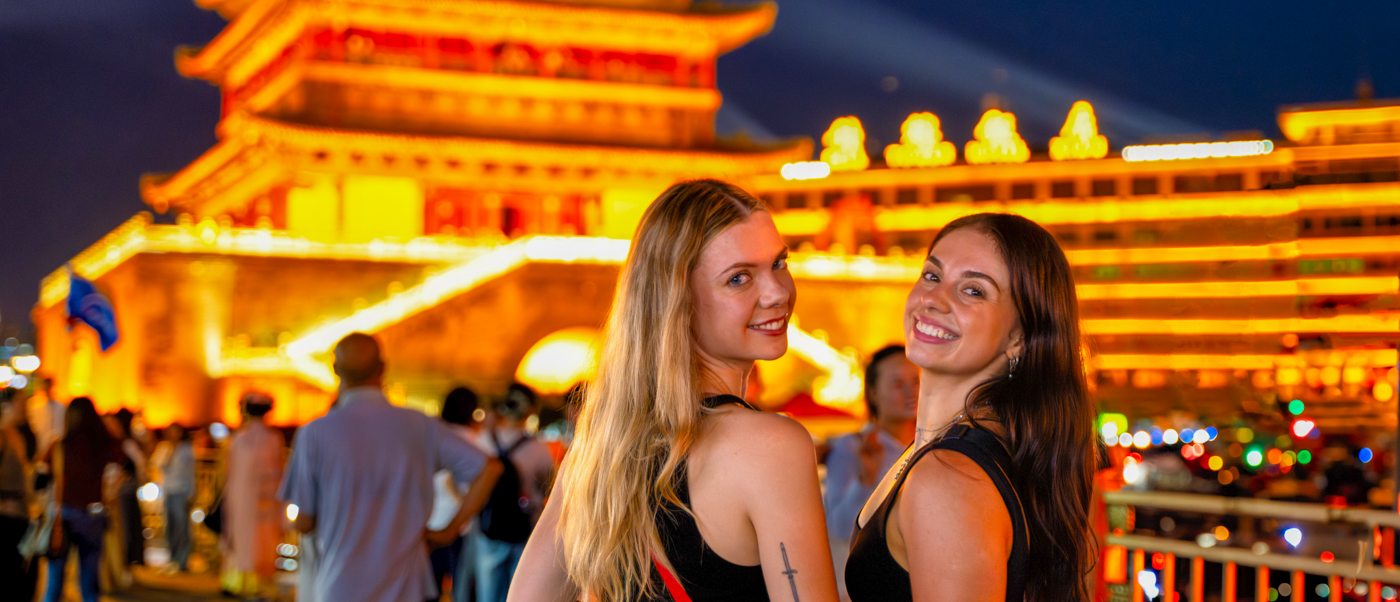 Two travellers smile in front of a brightly lit traditional tower on a busy evening street.